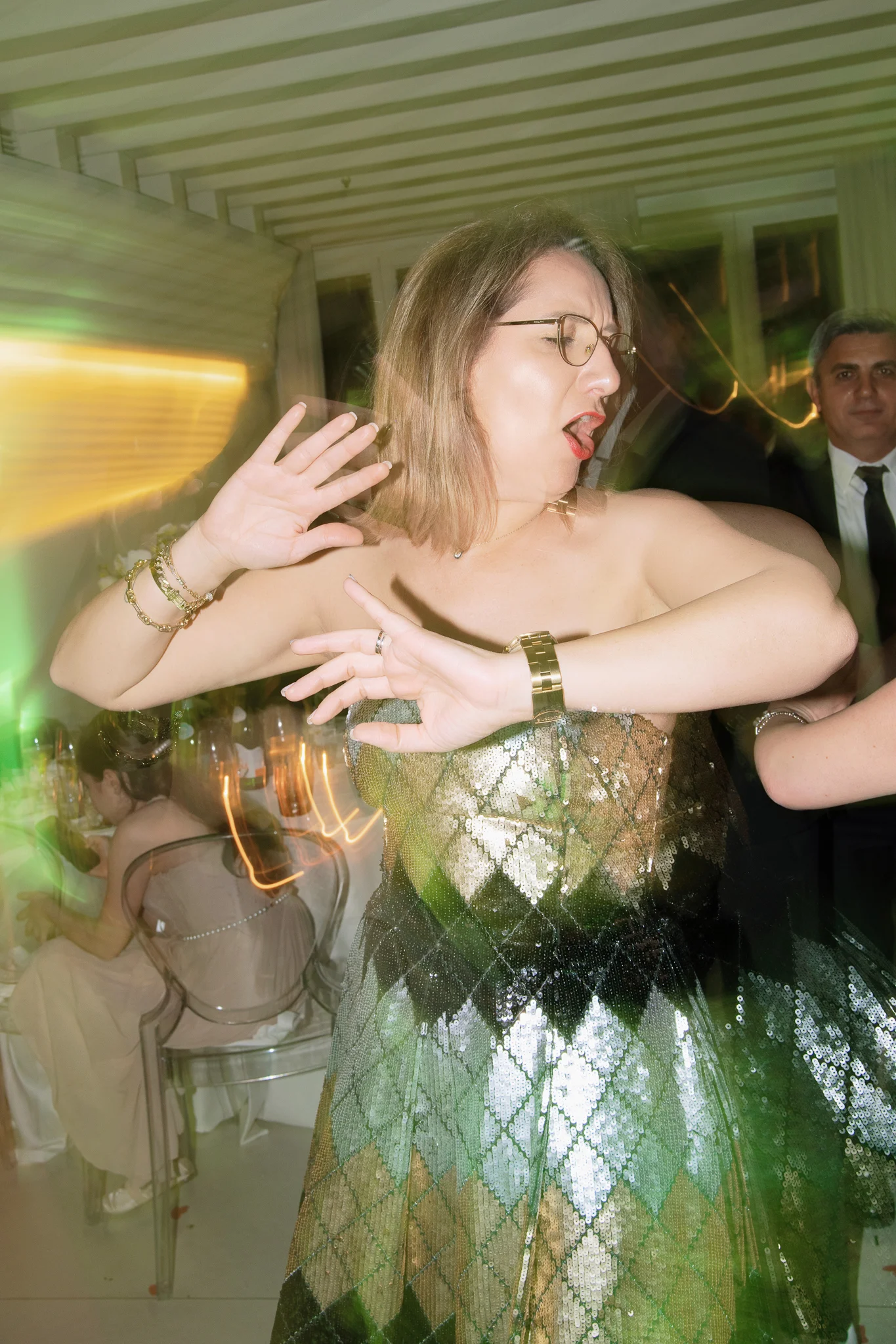 A female guest in a sparkly green and gold dress dances with her hands up at a lively wedding party.