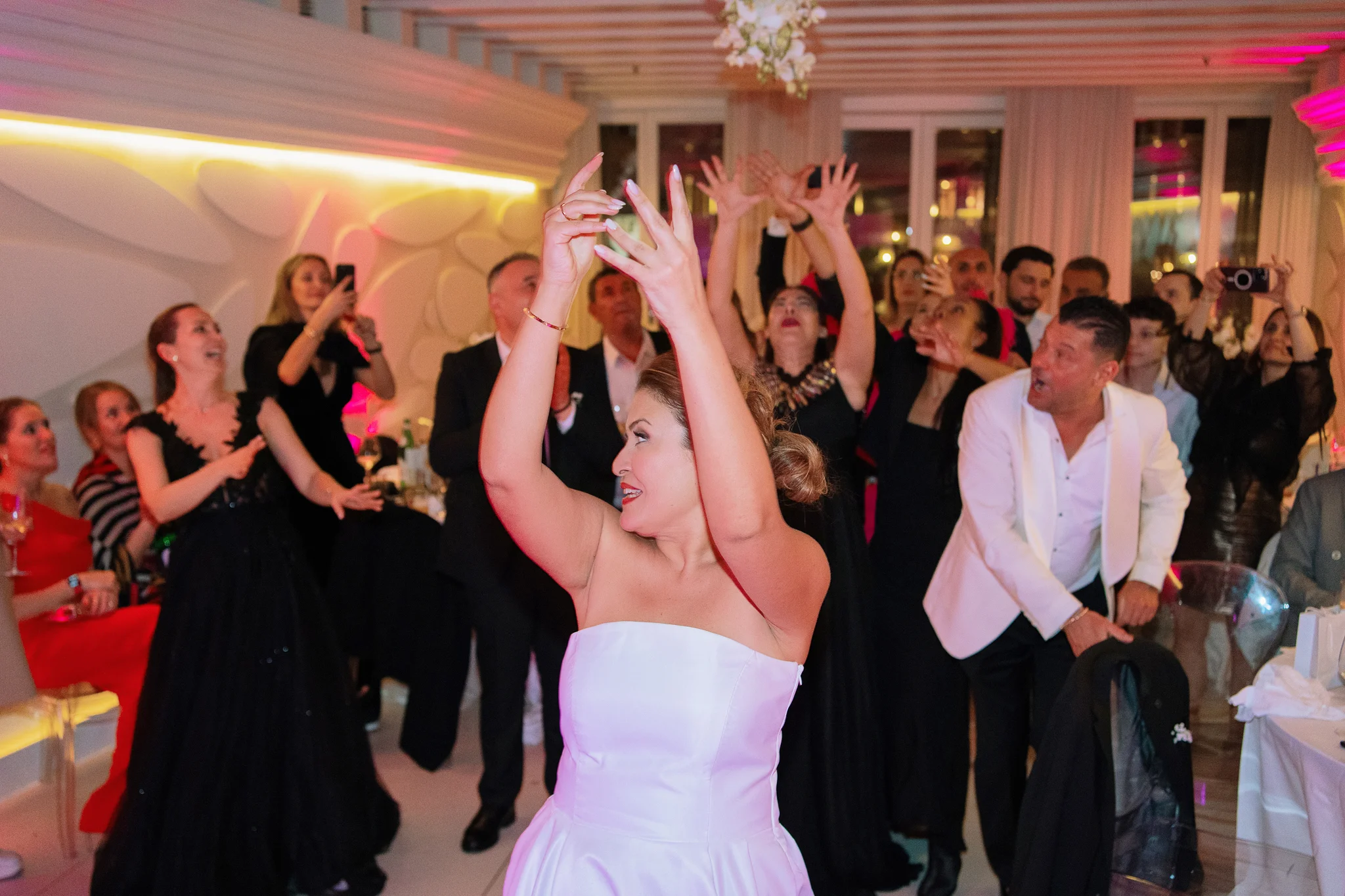 A bride prepares to toss her wedding bouquet to a group of excited guests waiting with hands up.