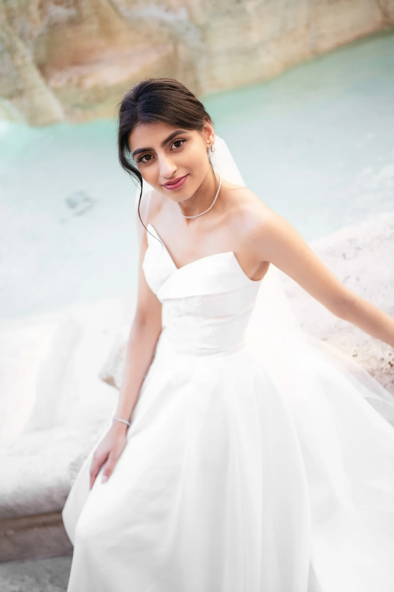 A beautiful bride in a white gown and veil sits by the turquoise water of a fountain.