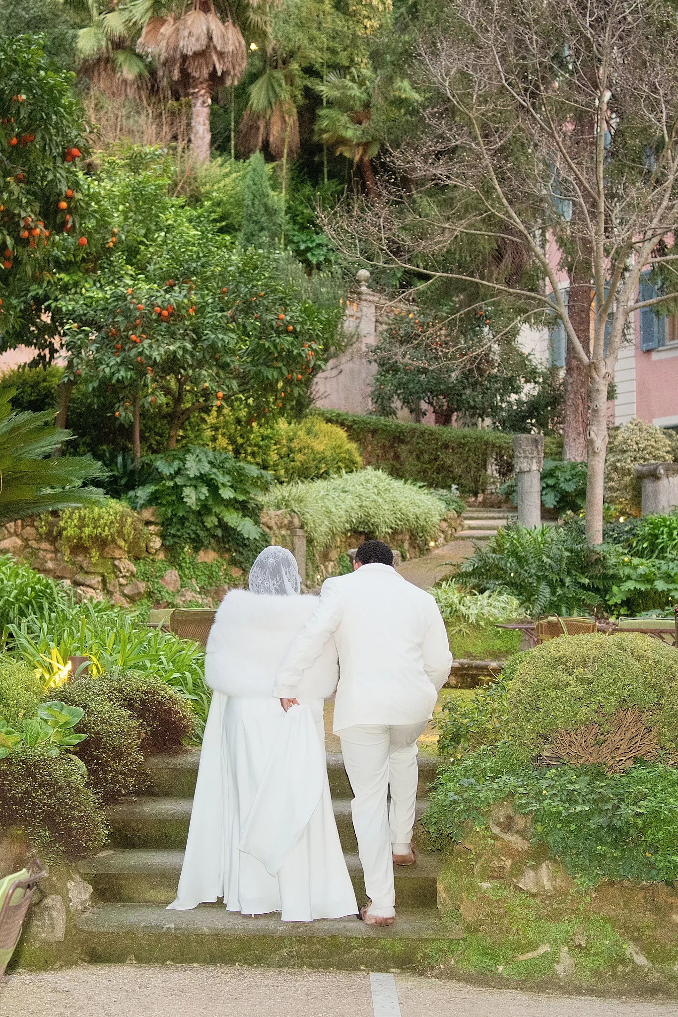 A couple dressed in white walks up stone steps together in a lush, green garden.