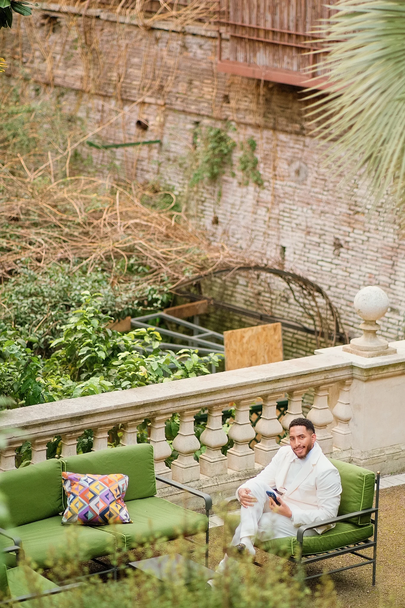 A stylish groom in a white suit relaxes on a green sofa on a garden terrace.