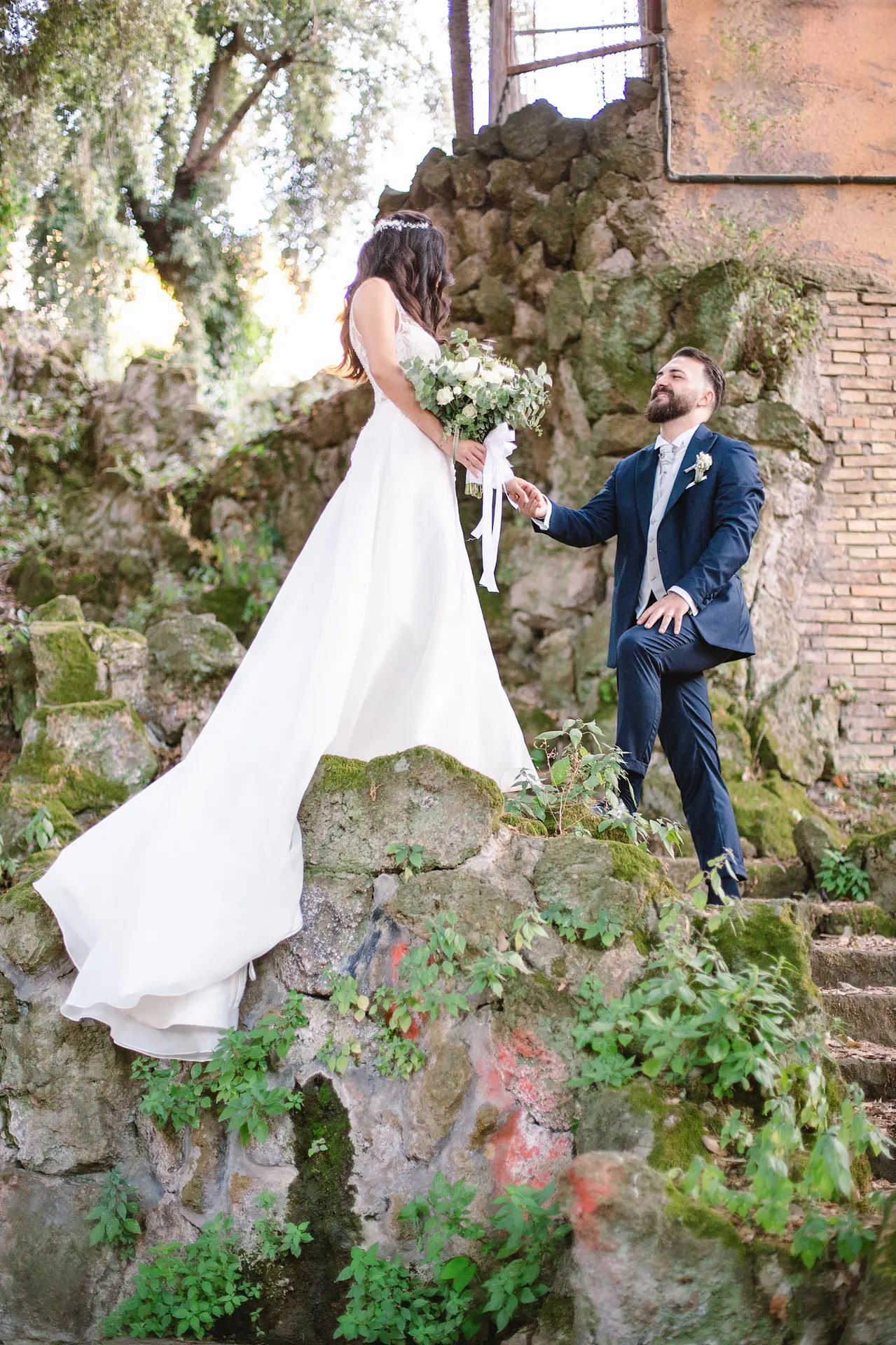 A groom on one knee on mossy stone steps, taking his bride's hand.