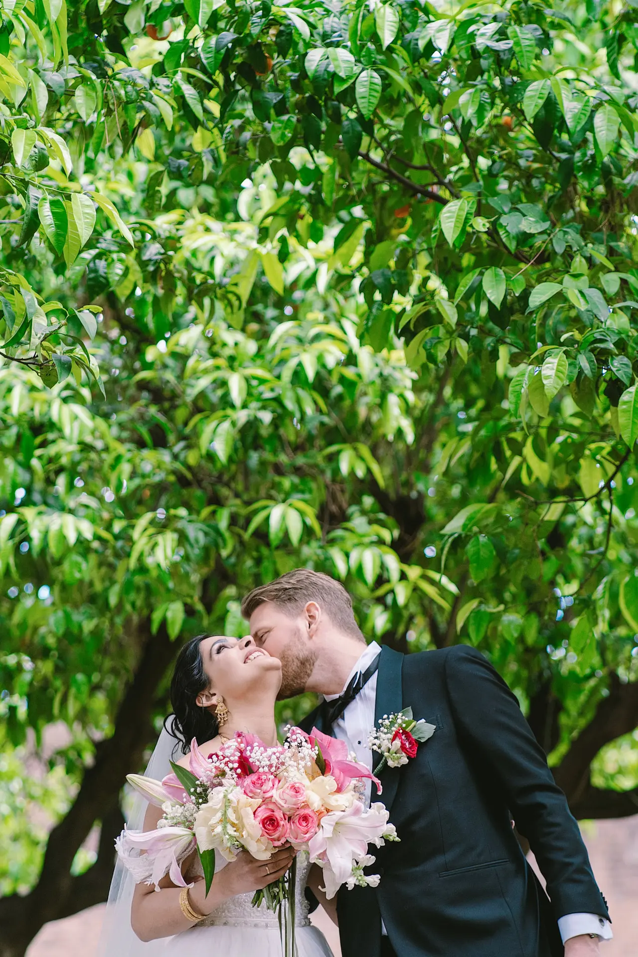 A groom kisses his smiling bride's cheek under a canopy of lush green leaves.