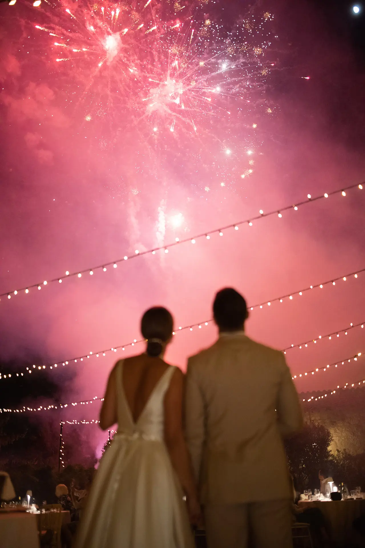 A newlywed couple holds hands while watching a spectacular pink fireworks display.