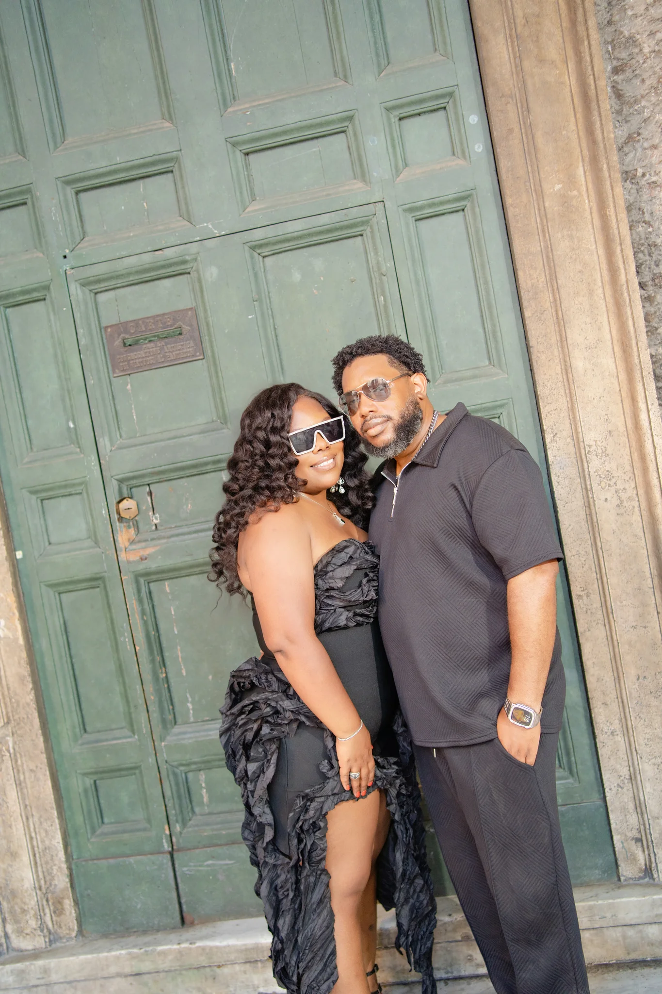 A fashionable couple wearing stylish black outfits and sunglasses pose together before a vintage green door.