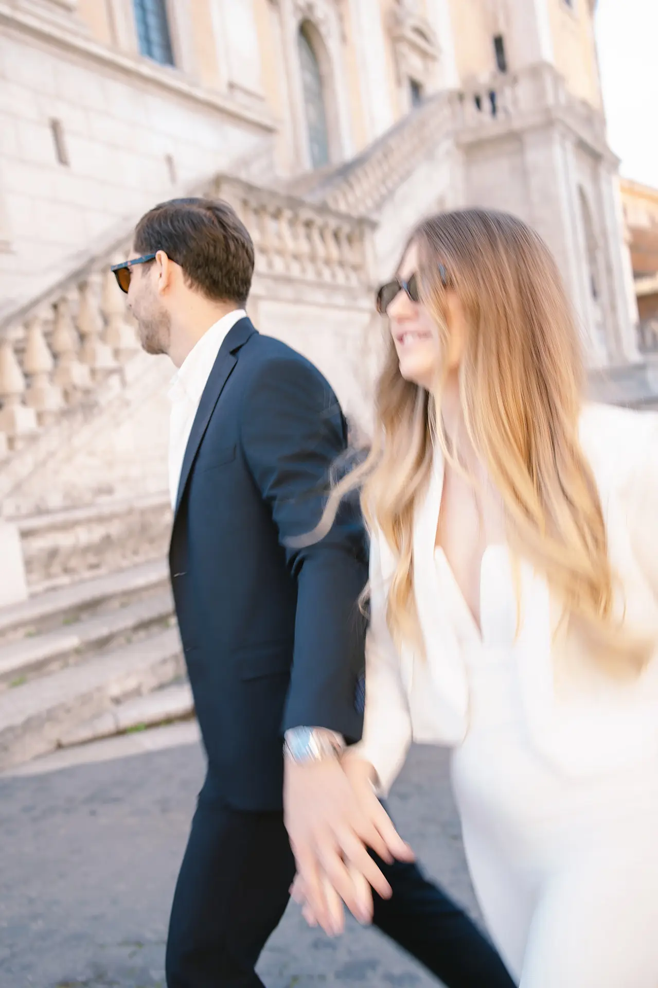 A dynamic motion blur captures a couple in sunglasses as they walk hand-in-hand through Roman streets.