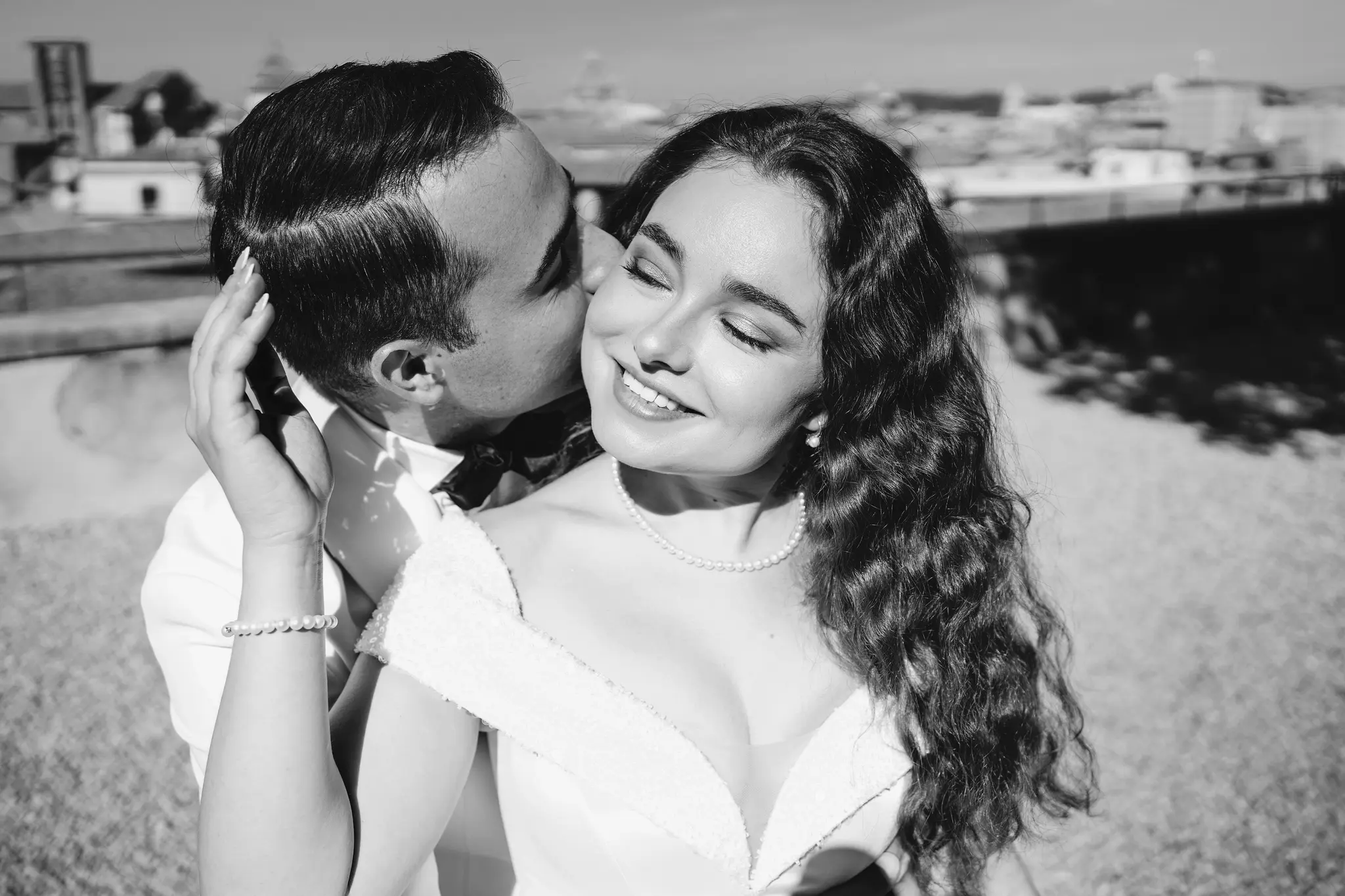 A black-and-white close-up of a man in a suit kissing a smiling bride on her cheek outdoors.