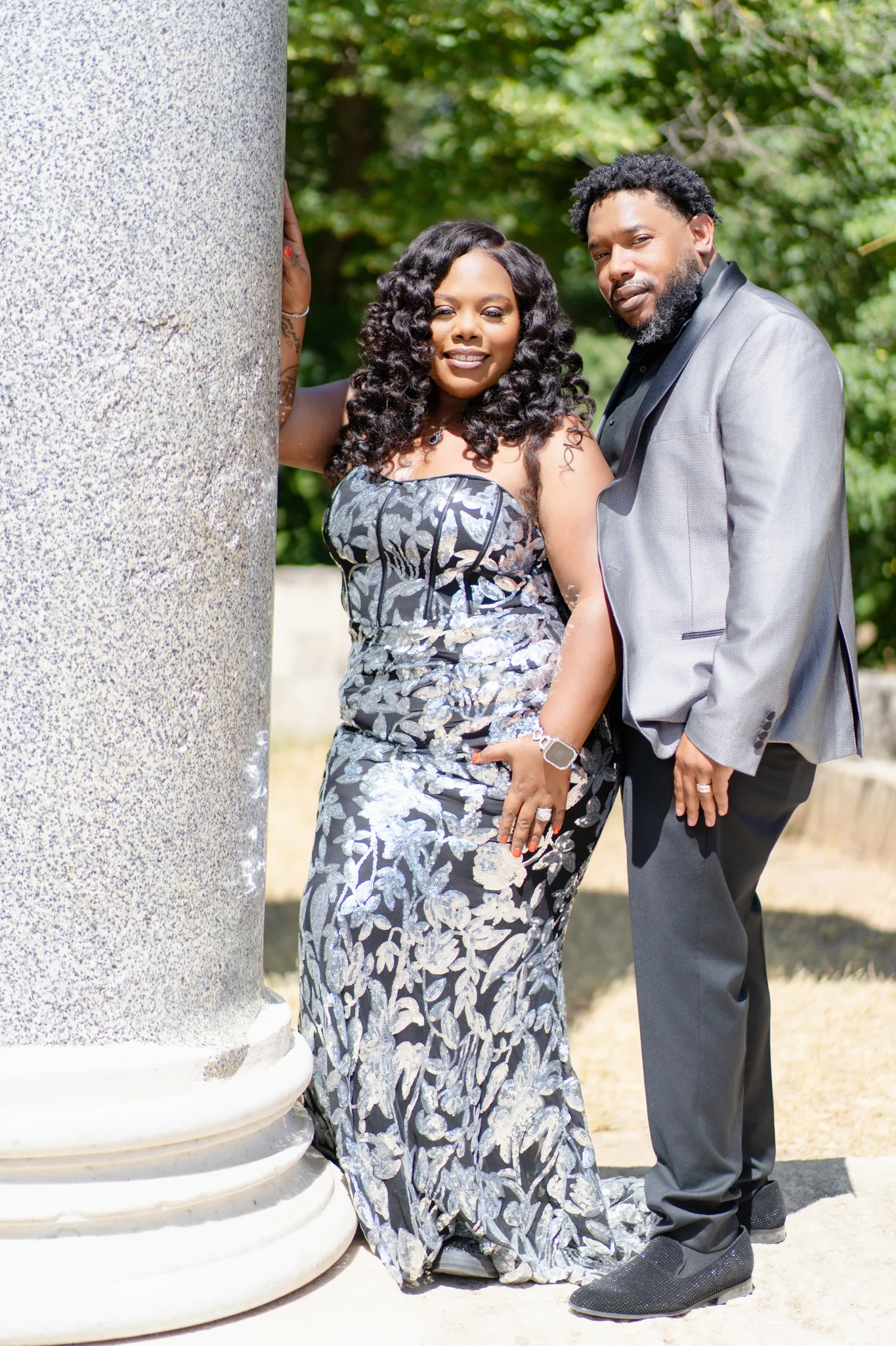 A smiling couple poses next to a large stone column; she wears a floral gown, and he wears a grey suit.