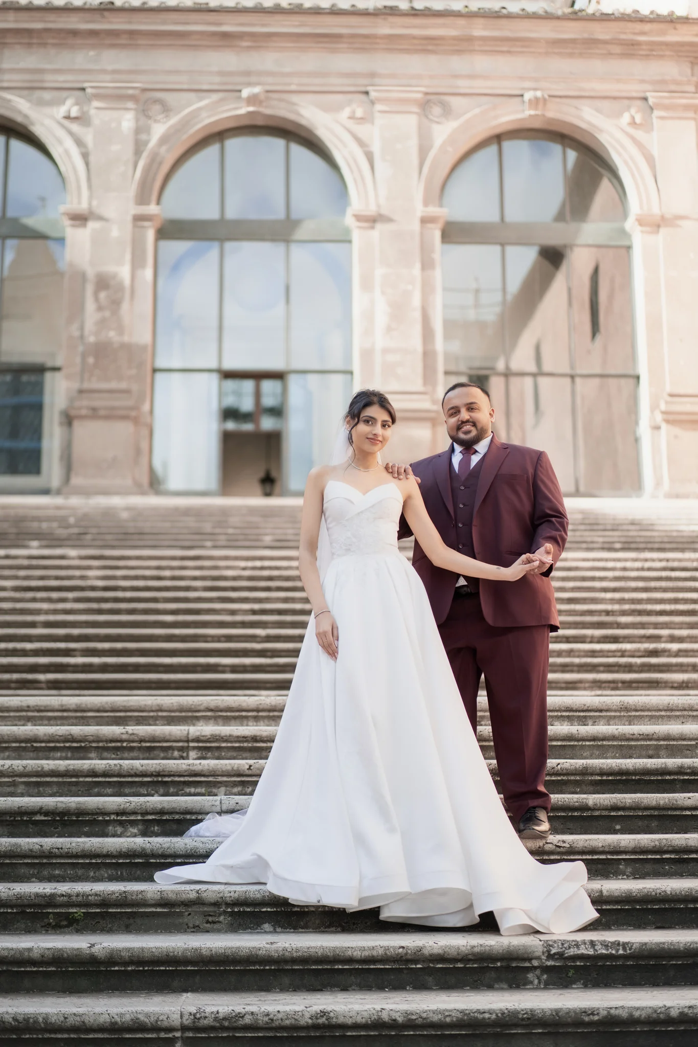 A couple in formal wear, a white gown and a maroon suit, pose holding hands on a grand stone staircase.