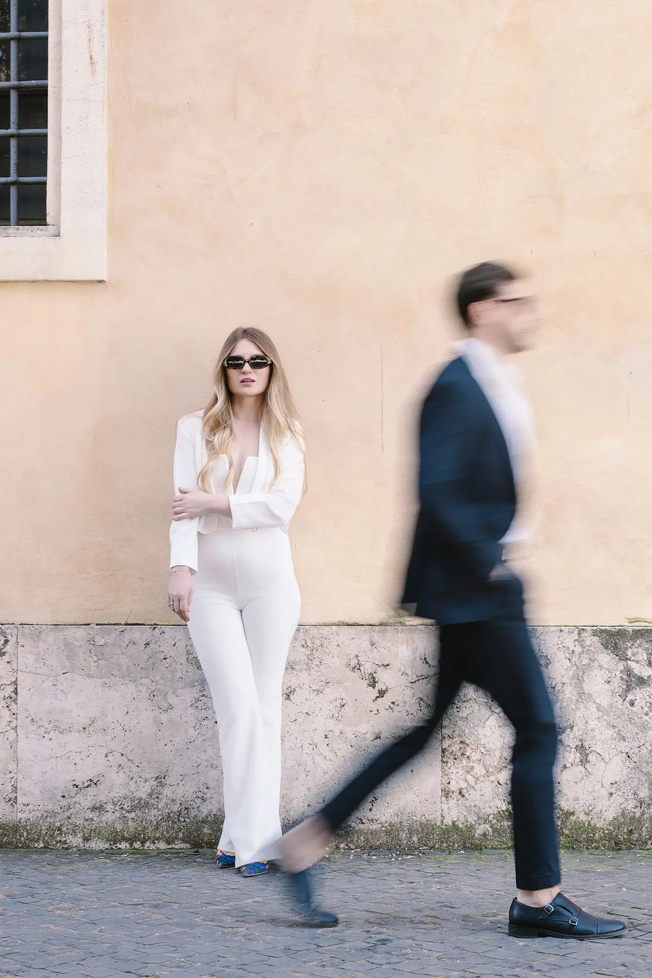 A woman in a white jumpsuit and sunglasses leans on a wall as a man in a suit walks by in motion blur.