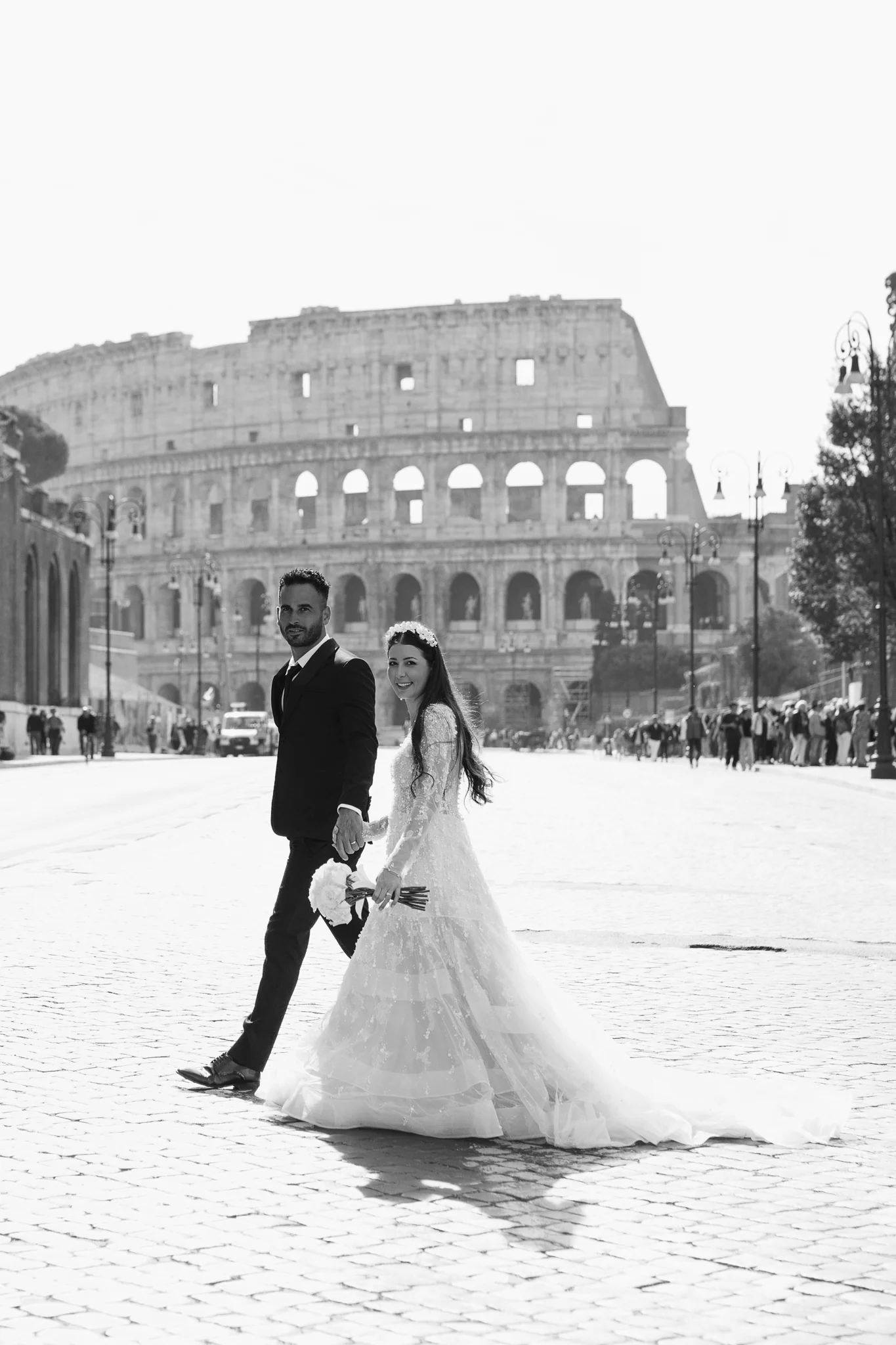 A couple in wedding attire walks hand-in-hand on a cobblestone street, with the Colosseum behind them.