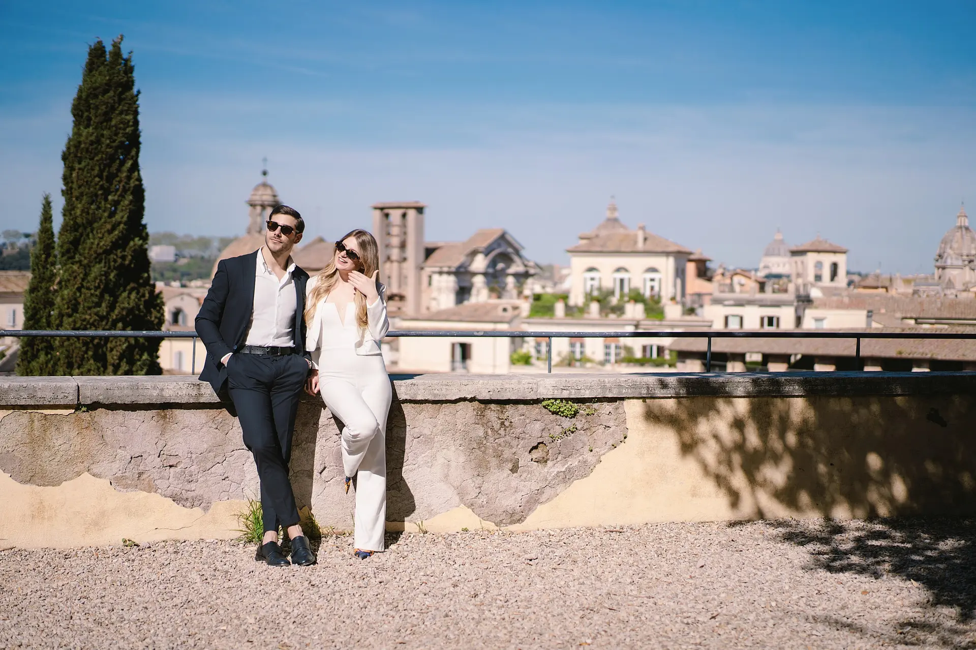 An engagement photographer in Rome captures a stylish couple leaning on a wall. The city skyline is behind them.
