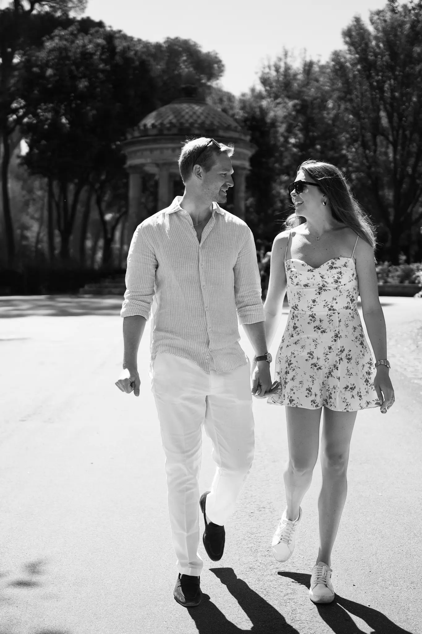 A black and white photo of a couple smiling at each other while walking hand-in-hand down a path in a park.