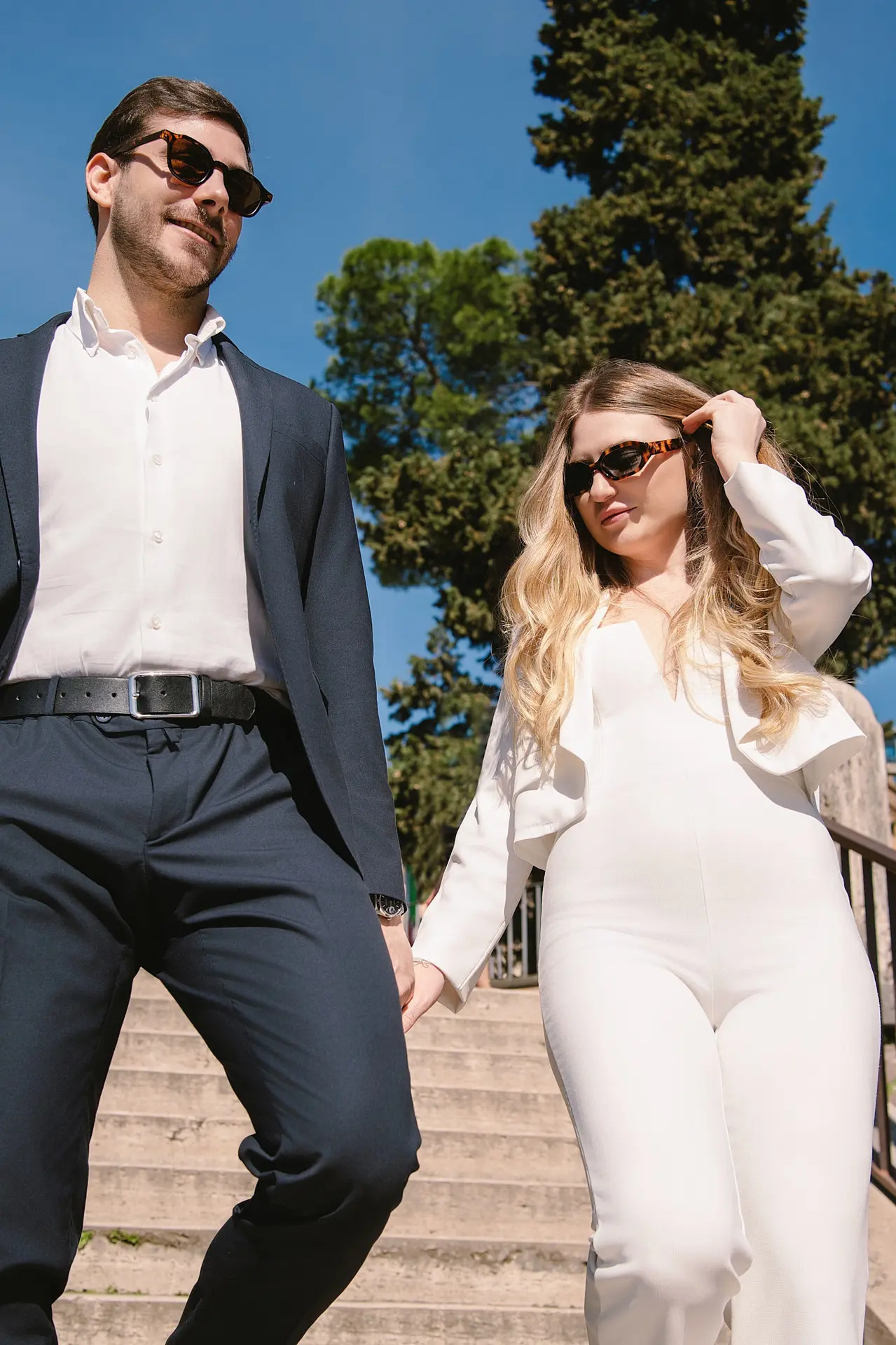 A low-angle shot of a stylish couple in sunglasses holding hands while walking down outdoor stone steps.