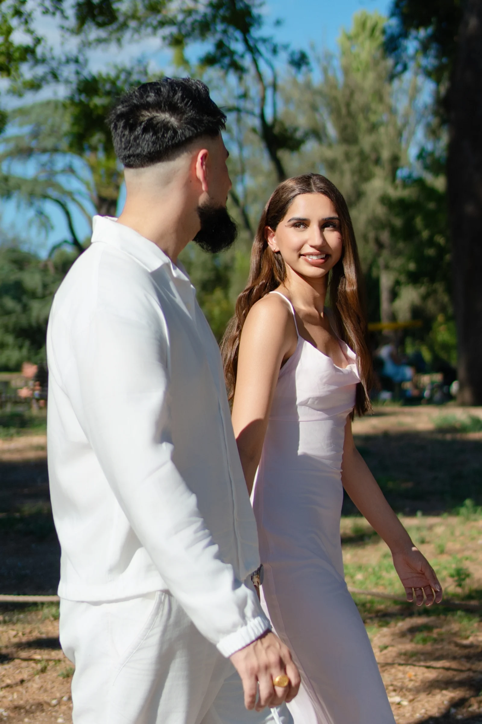 A woman in a pink dress walks hand-in-hand with a man. She turns her head to smile directly at the camera.