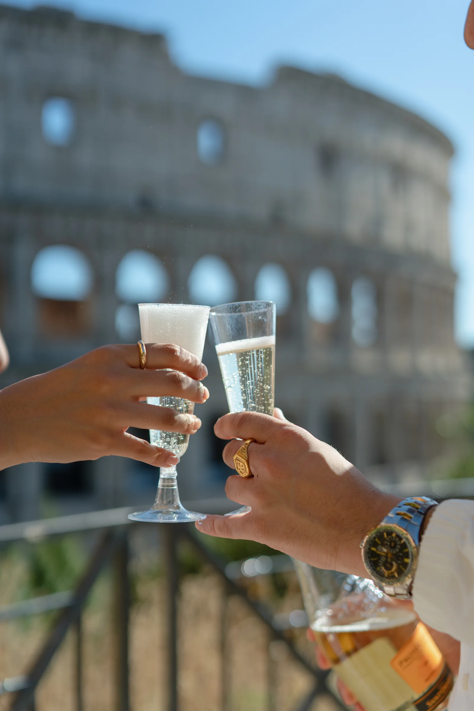 A close-up of two people toasting with champagne glasses. The Colosseum is visible in the background.