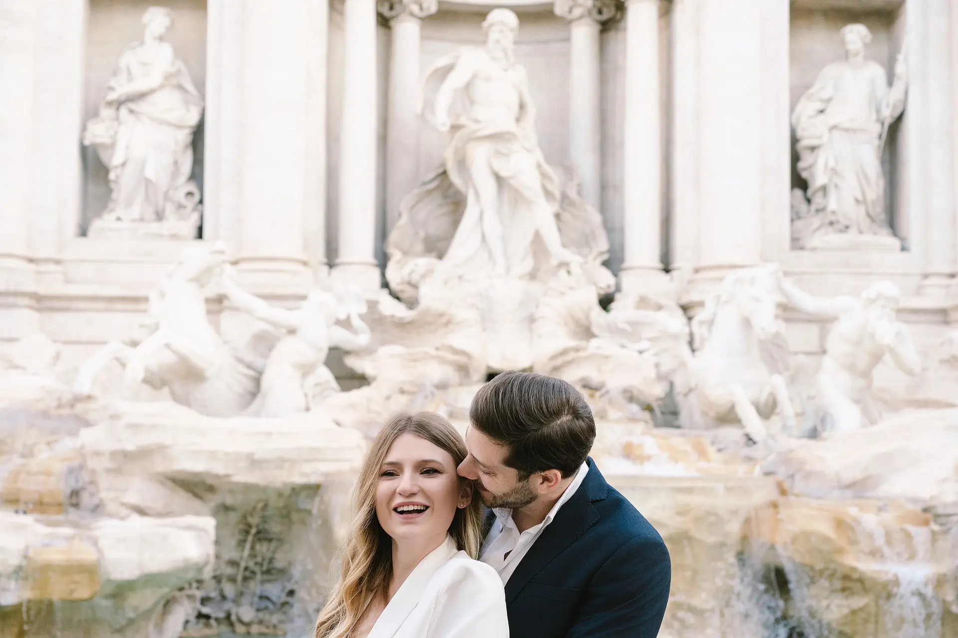 An engagement photographer in Rome captures a man kissing a smiling woman's cheek in front of the Trevi Fountain.