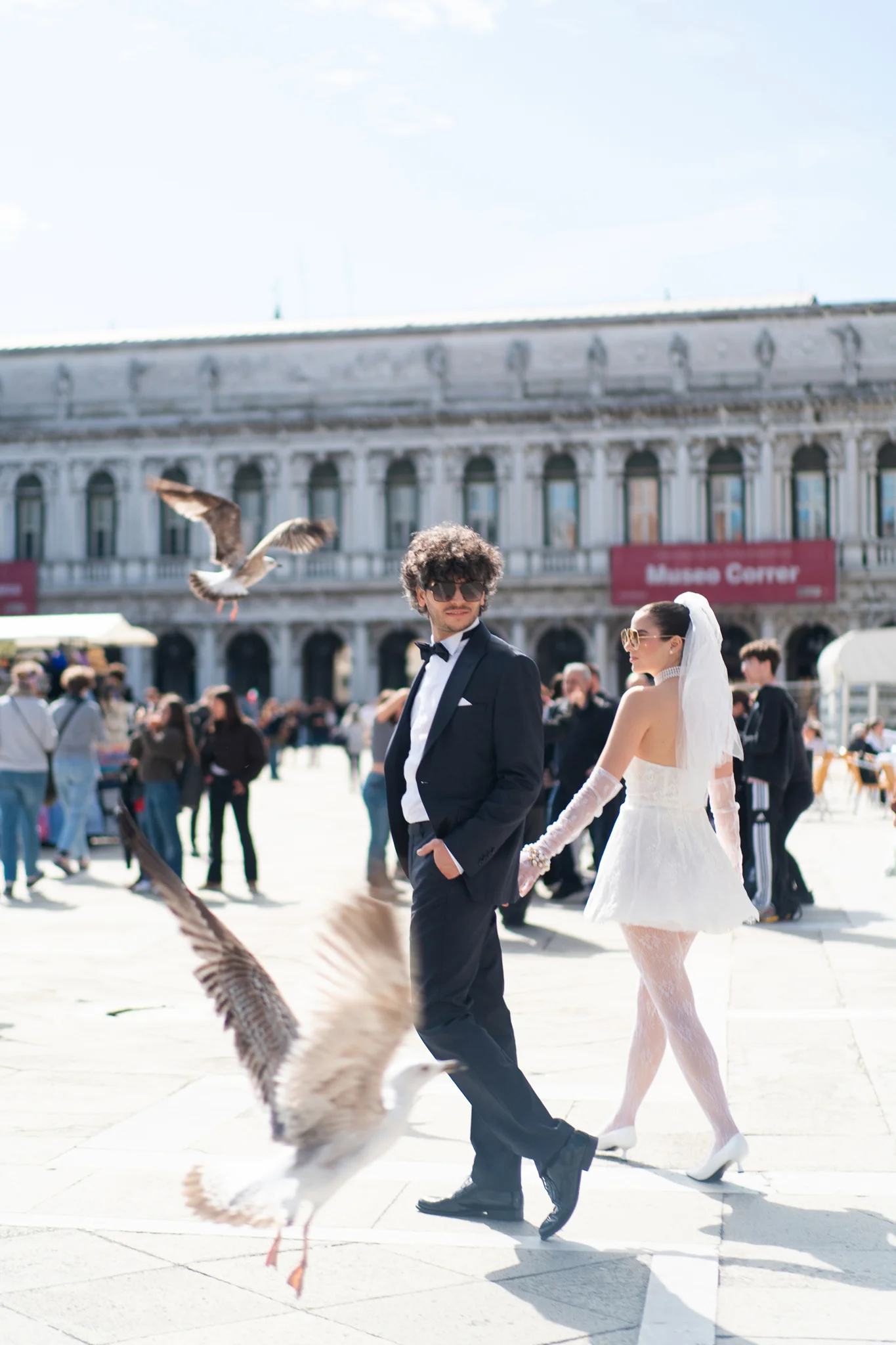 A newlywed couple in stylish attire walks through a busy Venice square with seagulls.