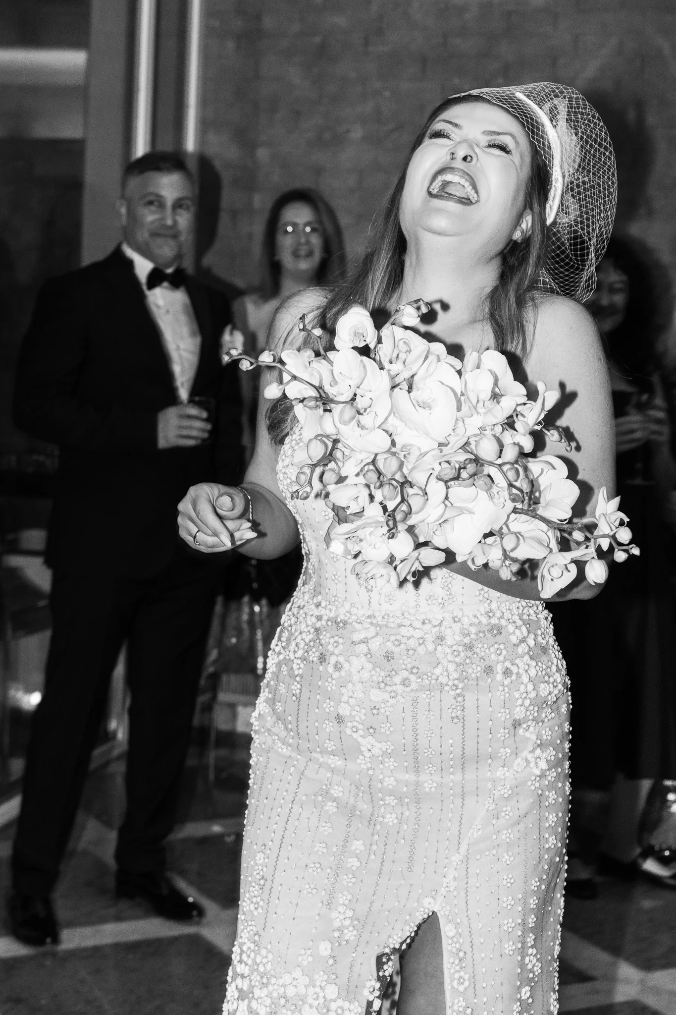 A black and white photo of a bride laughing heartily while holding her orchid bouquet.